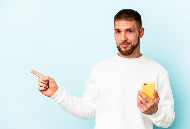 Young caucasian man holding mobile phone isolated on blue background smiling and pointing aside, showing something at blank space.