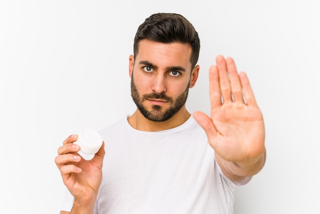 Young caucasian man holding a moisturizer isolated standing with outstretched hand showing stop sign, preventing you.