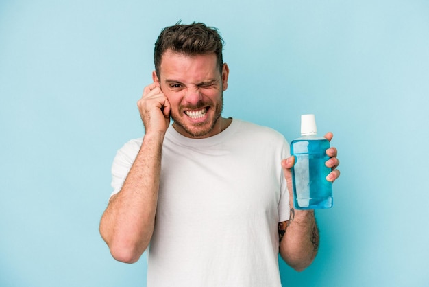 Young caucasian man holding mouthwash isolated on blue background covering ears with hands.