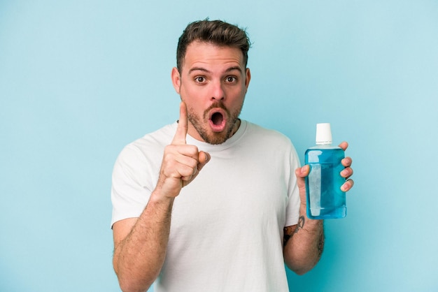 Young caucasian man holding mouthwash isolated on blue background having an idea, inspiration concept.