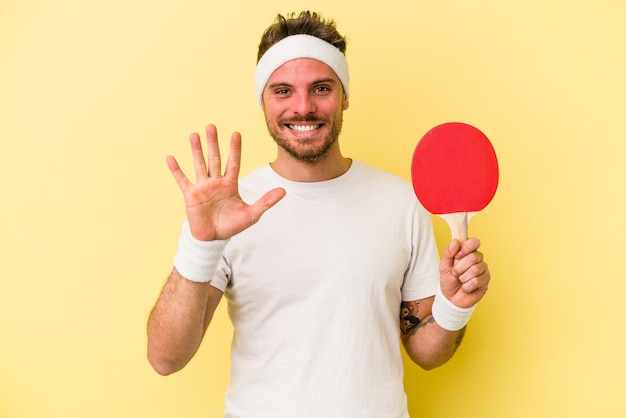 Young caucasian man holding ping pong racket isolated on yellow background smiling cheerful showing number five with fingers.