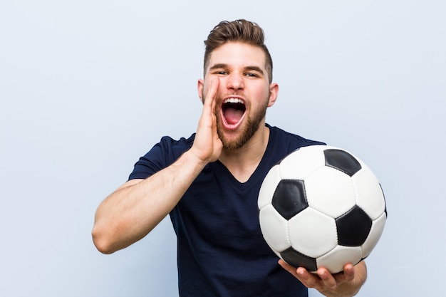 Young caucasian man holding a soccer ball shouting excited to front.