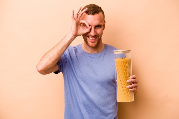 Young caucasian man holding spaghettis jar isolated on beige background excited keeping ok gesture on eye