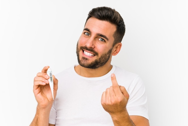 Young caucasian man holding a thermometer isolated Young caucasian man holding  a pointing with finger at you as if inviting come closer.