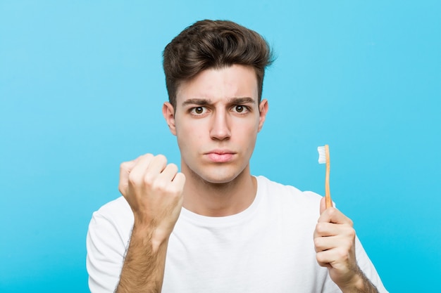 Young caucasian man holding a toothbrush Young indian woman wearing a pajamas and sleep mask isolated holding a pillow showing fist, aggressive facial expression.< mixto >