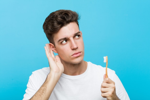 Young caucasian man holding a toothbrush Young indian woman wearing a pajamas and sleep mask isolated holding a pillow trying to listening a gossip.< mixto >