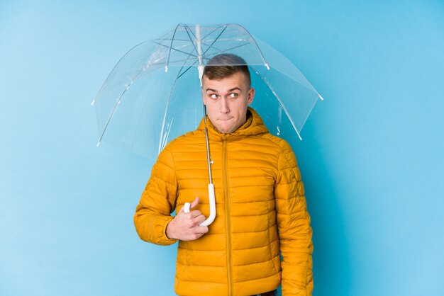 Young caucasian man holding an umbrella confused, feels doubtful and unsure.