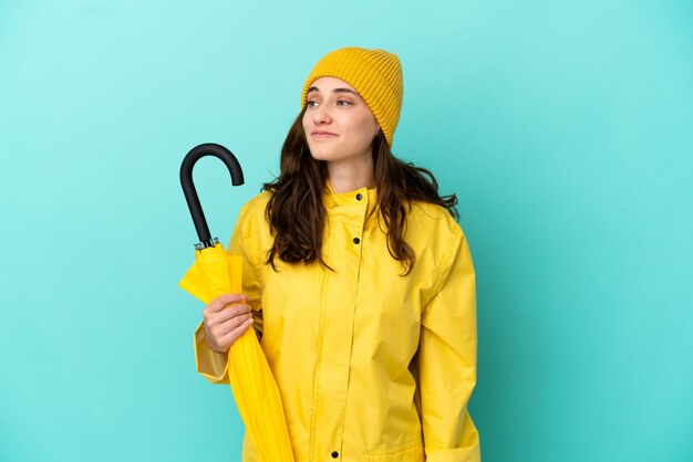 Young caucasian man holding an umbrella isolated on blue background looking to the side