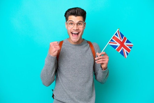 Young caucasian man holding an United Kingdom flag isolated on blue background celebrating a victory in winner position