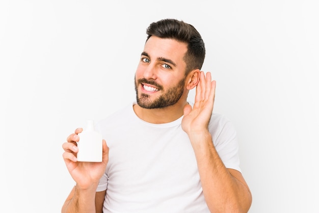 Young caucasian man holding a vitamins bottle isolated trying to listening a gossip.