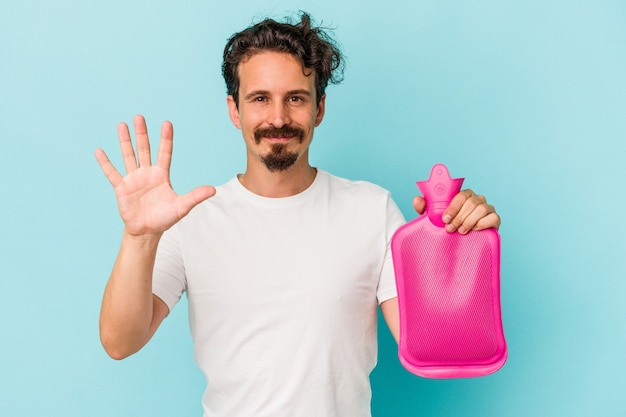 Young caucasian man holding a water bag isolated on blue background smiling cheerful showing number five with fingers.