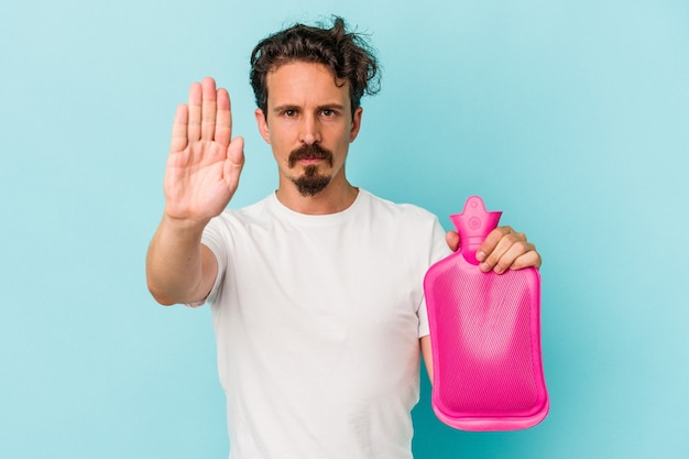 Young caucasian man holding a water bag isolated on blue background standing with outstretched hand showing stop sign, preventing you.