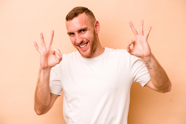 Young caucasian man isolated on beige background cheerful and confident showing ok gesture