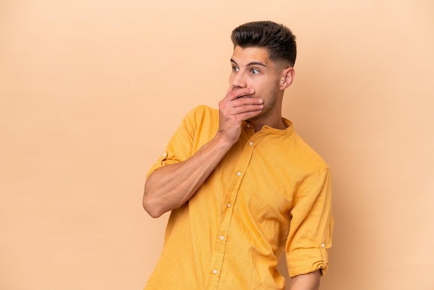 Young caucasian man isolated on beige background doing surprise gesture while looking to the side