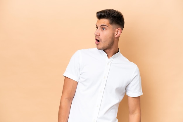 Young caucasian man isolated on beige background doing surprise gesture while looking to the side