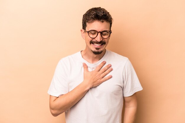 Young caucasian man isolated on beige background laughs out loudly keeping hand on chest.