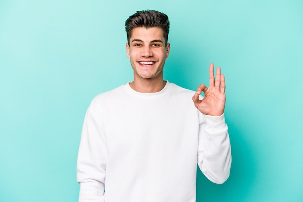 Young caucasian man isolated on blue background cheerful and confident showing ok gesture