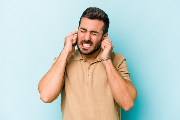 Young caucasian man isolated on blue background covering ears with fingers stressed and desperate by a loudly ambient