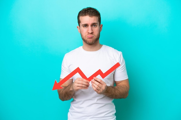 Young caucasian man isolated on blue background holding a downward arrow and with sad expression