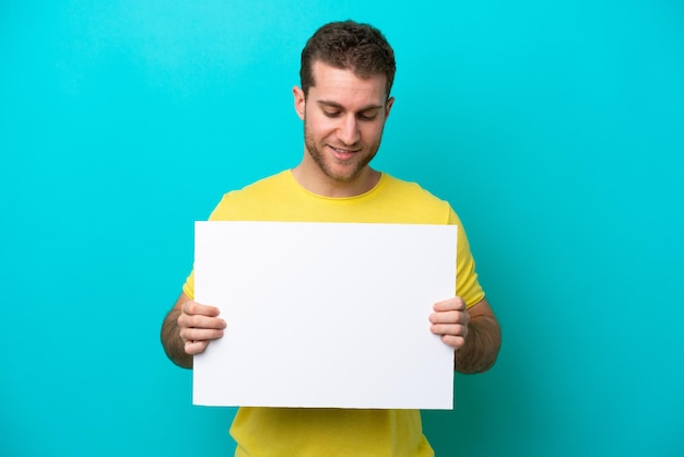 Young caucasian man isolated on blue background holding an empty placard and looking it