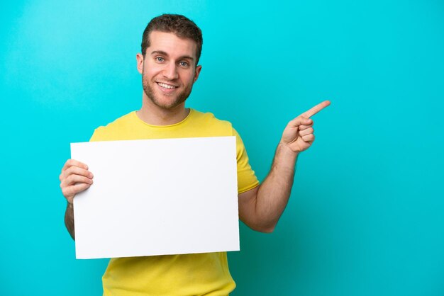 Young caucasian man isolated on blue background holding an empty placard and pointing side