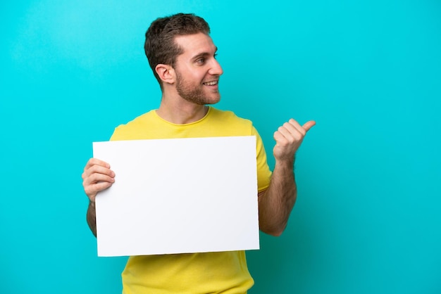 Young caucasian man isolated on blue background holding an empty placard and pointing side