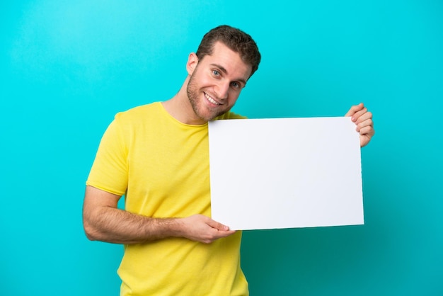 Young caucasian man isolated on blue background holding an empty placard with happy expression