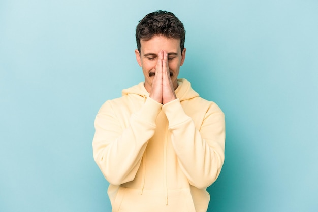 Young caucasian man isolated on blue background holding hands in pray near mouth, feels confident.