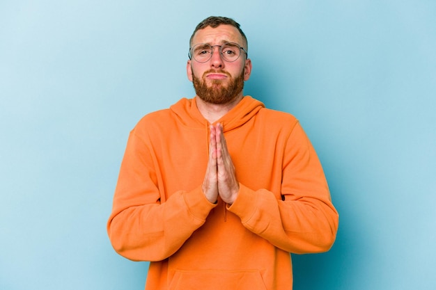 Photo young caucasian man isolated on blue background holding hands in pray near mouth, feels confident.