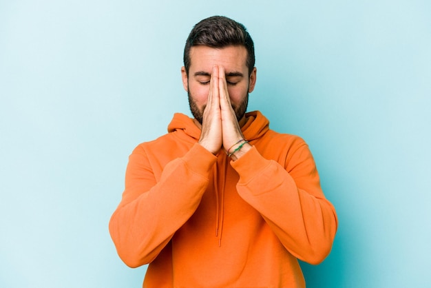 Young caucasian man isolated on blue background holding hands in pray near mouth feels confident