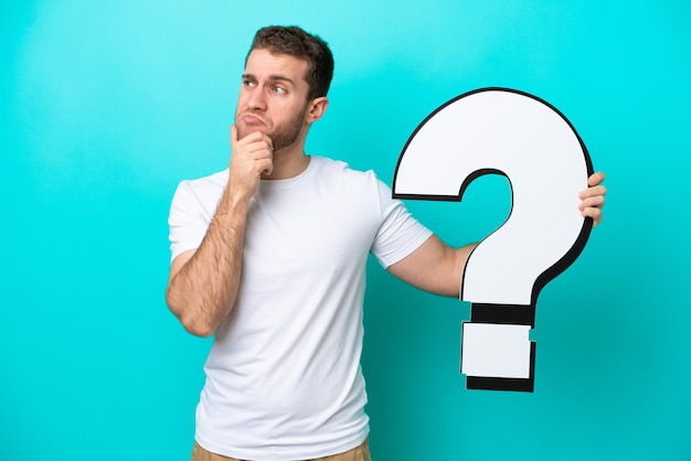 Young caucasian man isolated on blue background holding a question mark icon and having doubts