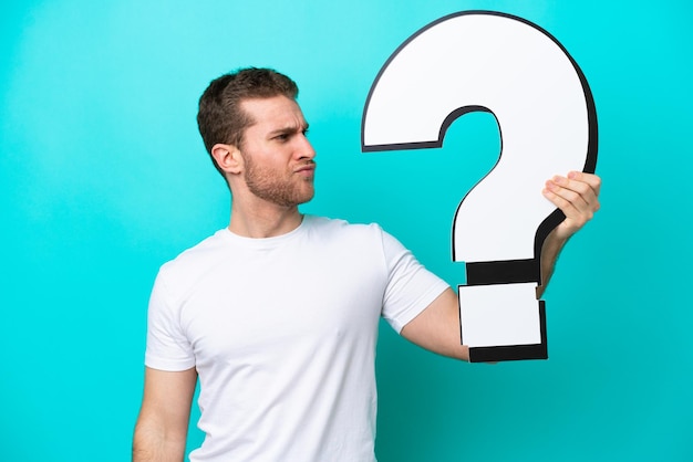 Young caucasian man isolated on blue background holding a question mark icon and having doubts