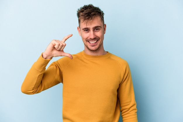 Young caucasian man isolated on blue background holding something little with forefingers, smiling and confident.