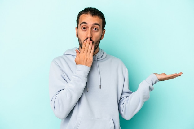 Young caucasian man isolated on blue background impressed holding copy space on palm.