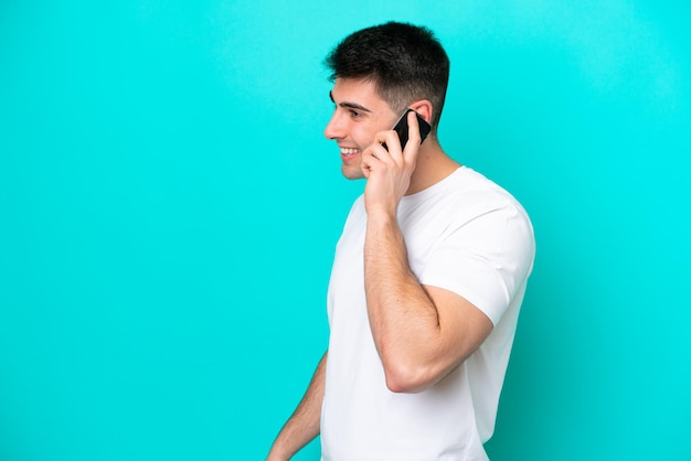 Young caucasian man isolated on blue background keeping a conversation with the mobile phone with someone