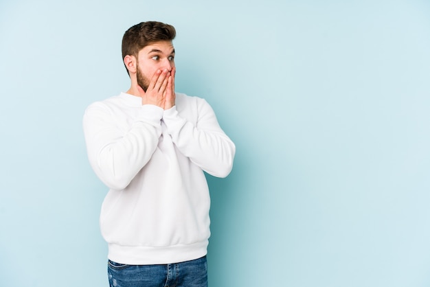 Young caucasian man isolated on blue background laughing about something, covering mouth with hands.