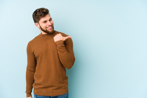 Young caucasian man isolated on blue background points with thumb finger away, laughing and carefree.