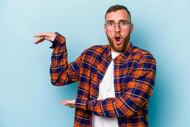 Young caucasian man isolated on blue background shocked and amazed holding a copy space between hands.