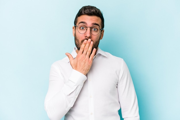 Young caucasian man isolated on blue background shocked covering mouth with hands anxious to discover something new