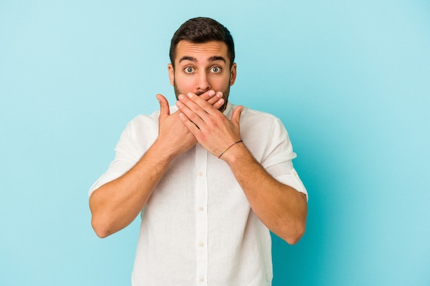 Young caucasian man isolated on blue background shocked covering mouth with hands.