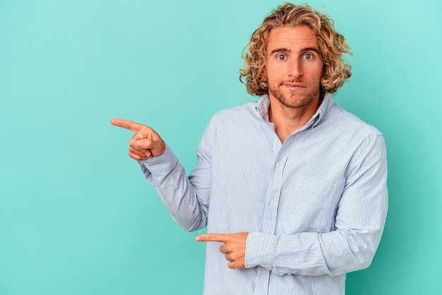 Young caucasian man isolated on blue background shocked pointing with index fingers to a copy space.