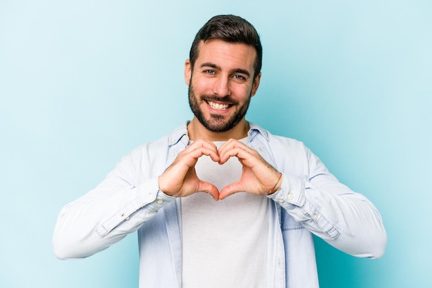 Young caucasian man isolated on blue background smiling and showing a heart shape with hands