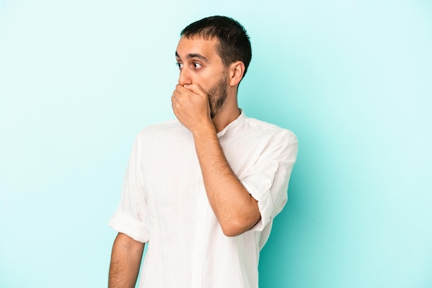 Young caucasian man isolated on blue background thoughtful looking to a copy space covering mouth with hand