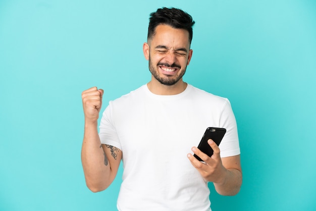 Young caucasian man isolated on blue background using mobile phone and doing victory gesture