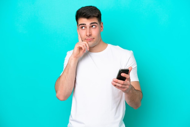 Young caucasian man isolated on blue background using mobile phone and thinking