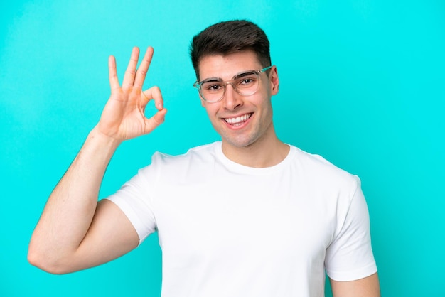 Young caucasian man isolated on blue background With glasses and doing OK sign