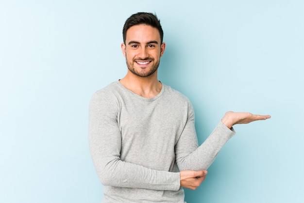 Young caucasian man isolated on blue showing a copy space on palm and holding another hand on waist.