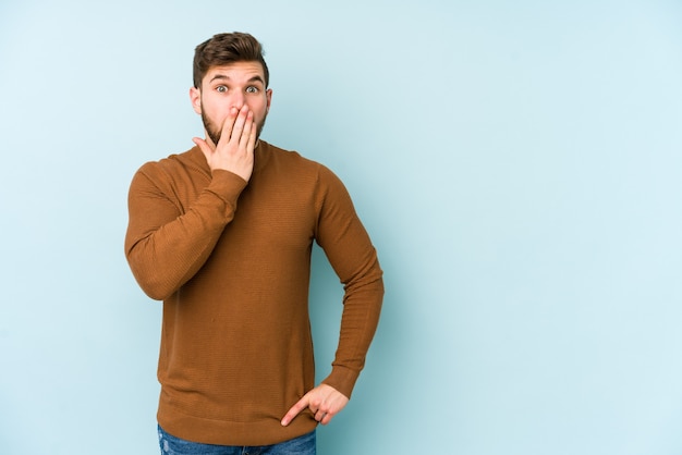 Young caucasian man isolated on blue space shocked, covering mouth with hands, anxious to discover something new.