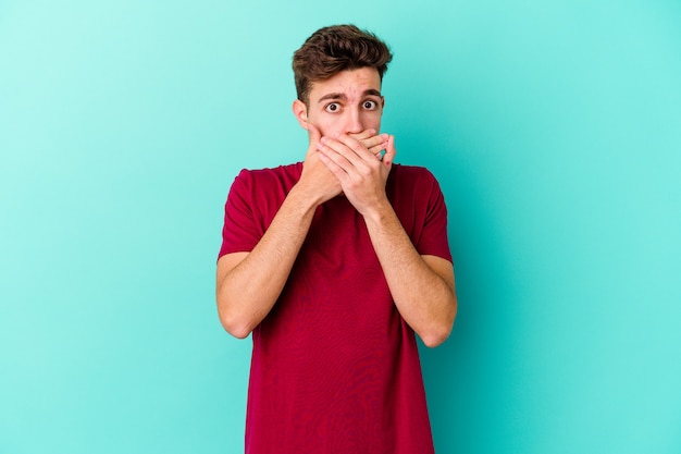 Young caucasian man isolated on blue wall shocked covering mouth with hands.