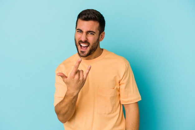 Young caucasian man isolated on blue wall showing rock gesture with fingers